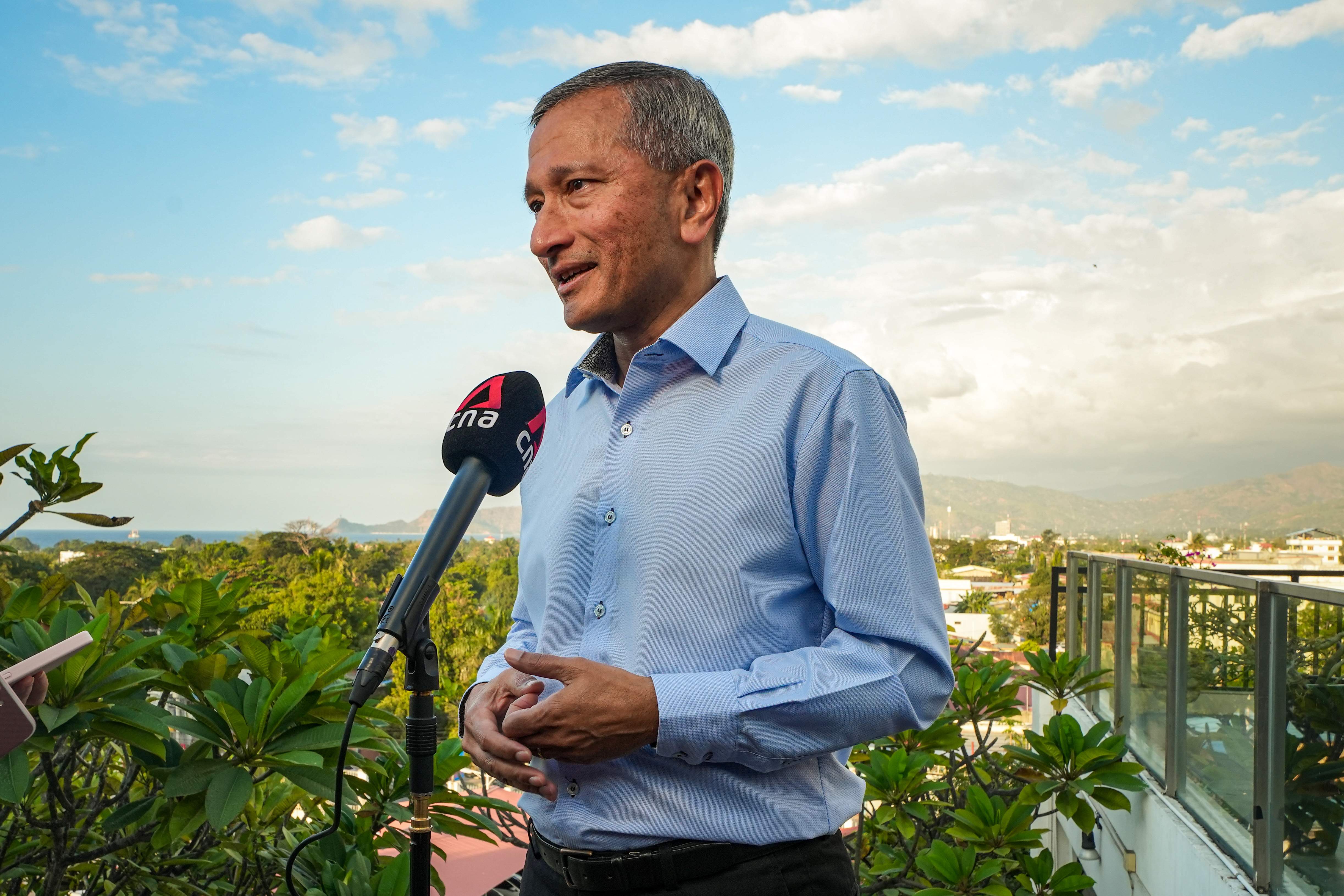 Man in light blue shirt speaks into CNA microphone outdoors, with skyline in background.